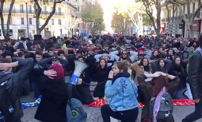 High school students marching in Marseille stop in the city centre to take up the mass knelt stress position in tribute/solidarity to the students of the Mates-la-Jolie Lycee whom were detained by police for hours in this manner.  It has become a symbolic gesture of defiance to Macron and violent state repression for the student movement and others. Photo: Video still, Twitter/laprovence