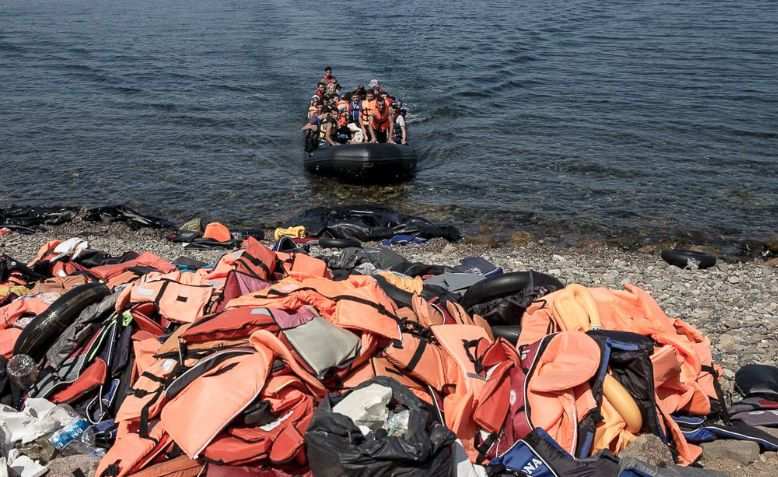 Migrants and refugees arrive by dinghy behind a huge pile of life vests after crossing from Turkey